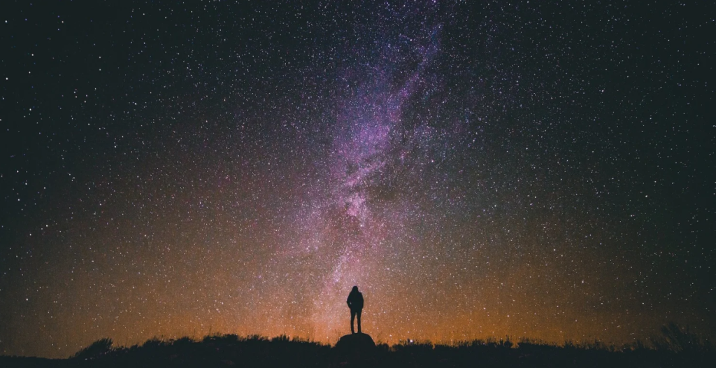 A person standing on a hill silhouetted against a star-filled night sky with the Milky Way above them.